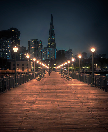 Woman walking on Pier 7 at night, with city downtown in the backdrop including the Transamerica Pyramid building, North Waterfront, Embarcadero, San Francisco, California, United States (July 2022)