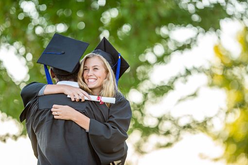 Two University graduates hug each other at the end of their ceremony, as they celebrate their success.  They are each wearing caps and gowns and holding their diplomas as they smile with pride.