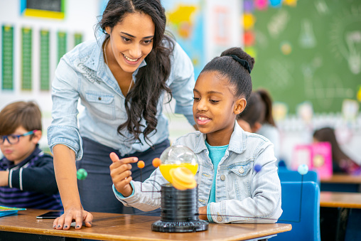 An elementary student sits at her desk with a model of the solar system out in front of her.  She is dressed casually and focused on the  activity as she explains how the system works to her teacher.