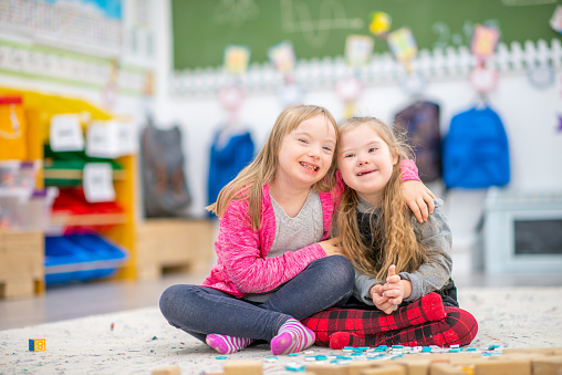 Two young elementary students with Down Syndrome, sit on the floor of their classroom as hey pose together for a portrait.  They are both dressed casually and are huddle in closely together.