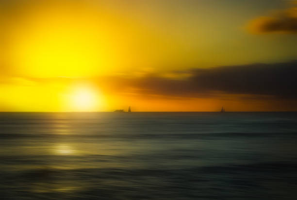 The image shows a sunset over calm ocean waters in Oahu, Hawaii, with sailboats scattered across the sea and an orange yellow glow in the sky.