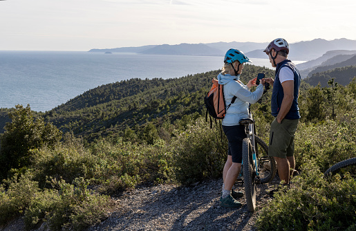 Mature mountain bikers pause on top of mountain and use mobile phone for direction, Liguria