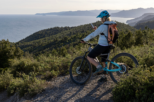 Mature e-mountain biker uses mobile phone on mountain top above sea, Liguria
