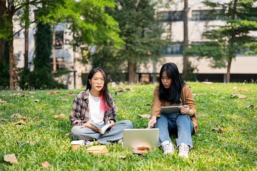 Two young Asian female college students are sitting on the grass in a park, using a laptop together, discussing something while studying or working on a project outdoors.