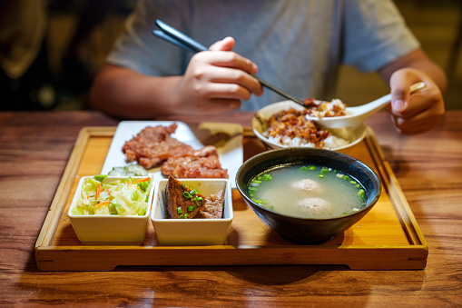 Close up shot of a young adult is seen enjoying a Taiwanese-style bento, a boxed meal consisting of braised pork rice, soup, and side dishes.