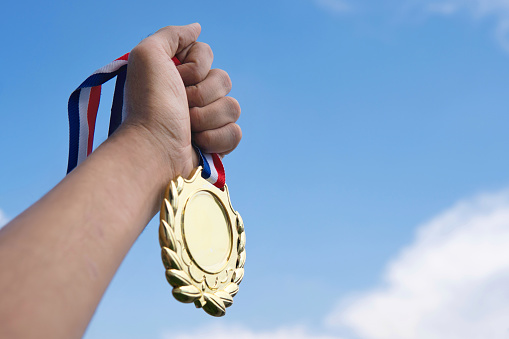 A hand triumphantly holds a gold medal aloft against a backdrop of clear blue sky. Copy space for text. Winner, achievement and success concept.
