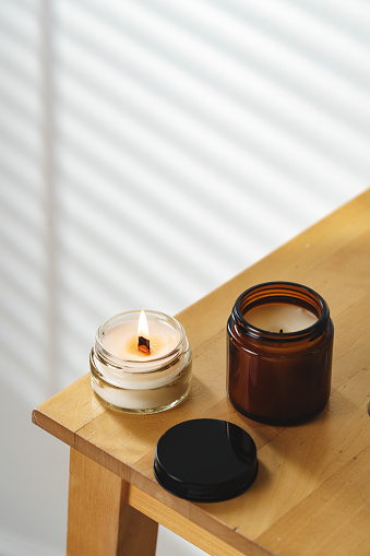 A peaceful scene is set with a gently flickering scented candle providing a warm glow on a wooden nightstand, suggesting a quiet, relaxed afternoon in a cozy room with soft natural light filtering through window blinds.