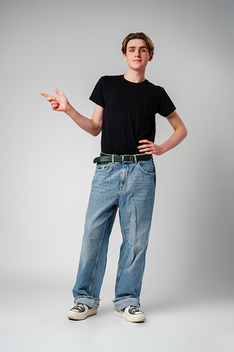 A young man with light brown hair stands confidently against a neutral gray background, wearing a plain black t-shirt paired with casual pants. He is gesturing to his side with an open hand as if presenting or explaining something.