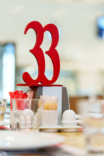 A bright red, bold number 33 stands prominently atop a restaurant table signifying its assignment amidst a blur of diners. The table includes condiment holders, cutlery, and menu holders suggestive of a casual dining environment, bustling with the energy of a lunchtime crowd.
