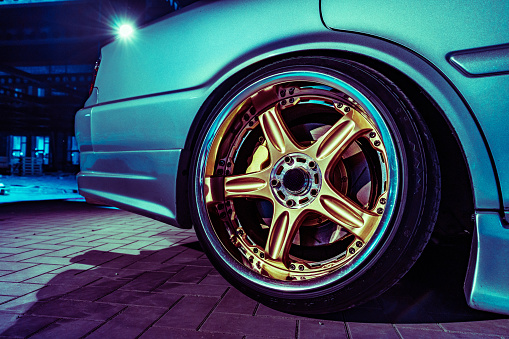 A close up shows the details of a golden alloy wheel on a sleek blue sports car, highlighting the shiny rim and tire tread in what appears to be a nighttime setting.