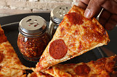 Close-up image of hand holding pepperoni pizza slice chosen from metal pizza pan, circular slices of pepperoni sausage, melted golden buffalo mozzarella cheese and rich tomato marinara sauce topping, chilli and herb shakers in background, elevated view