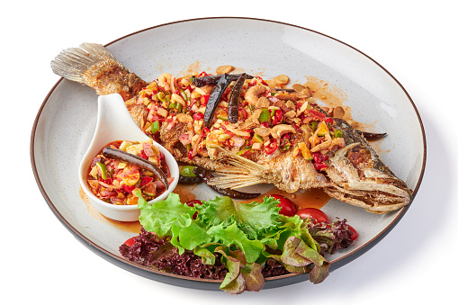 Deep fried fish with Thai spicy mixed herbs salad served with dipping sauce (sliced chili, lime, cashew nut, ginger, shallot) and fresh organic salad on plate isolated in white background