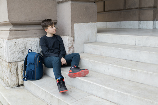 Schoolboy with backpack is sitting on staircase near the school building on first school day. Back to school. Education.