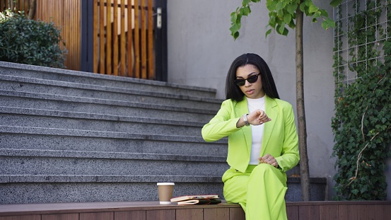 Portrait of American woman in green office sitting outside and checking time. Slow motion