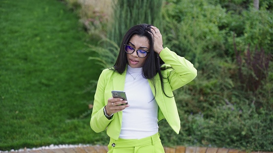 Female in green office clothes, holding smartphone, standing in park, and gets upset. Slow motion