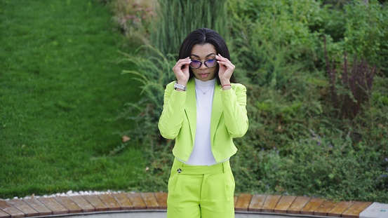 Woman in green office clothes, standing in park, and touching sunglasses. Slow motion