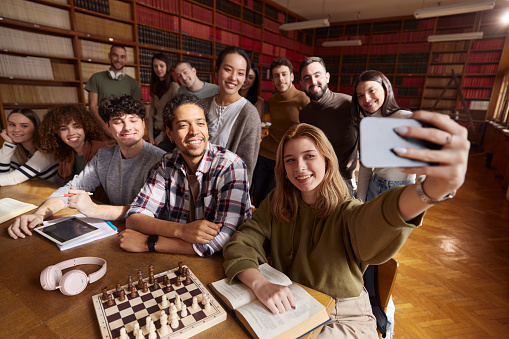 Large group of happy university students having fun while taking a selfie with cell phone in library.