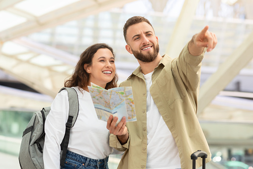 Travelling Concept. Happy Young Couple With City Map Standing Near Airport Building After Arrival To New Country, Cheerful Millennial Man And Woman Planning Travel Route, Man Pointing Away