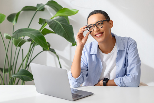 Positive millennial latin lady in glasses at table with computer, enjoy work and study, in white cafe, office interior. Communication remotely, app, gadget for business, freelance