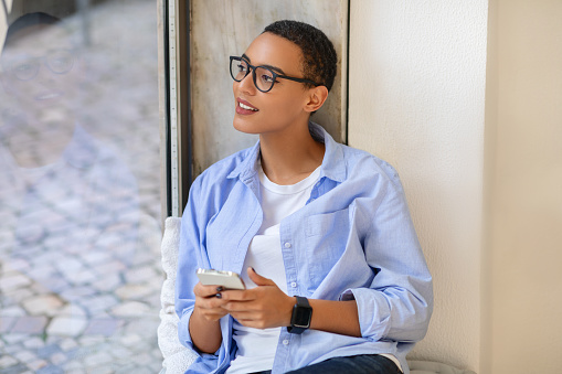 Pensive millennial latina lady in glasses, enjoy chat on mobile phone with smartwatch, look at free space, dream, think in cafe. Social networks and lifestyle, break alone and app