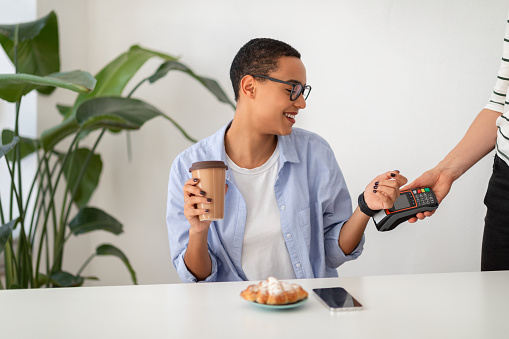 Cheerful young latin woman using smartwatch for payment machine, drink coffee with phone with empty screen on desk at white cafe. Modern contactless payment, relax indoor