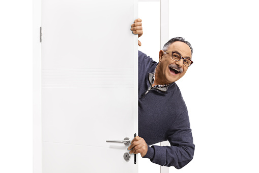 Cheerful mature man peeking from behind a white door isolated on white background