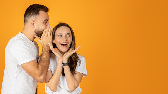 A glad european man whispers a secret into a woman's ear, both wearing white t-shirts, with the woman showing a surprised and delighted expression on an orange background