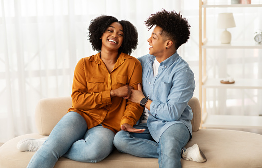 A laughing african american woman and a man looking at each other with affection while sitting closely on a couch, creating a sense of warmth and intimacy in a bright room