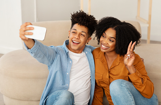 A cheerful young couple captures a joyful moment with a selfie, the man in a denim shirt flashing a bright smile as the woman in a burnt orange blouse poses with a peace sign