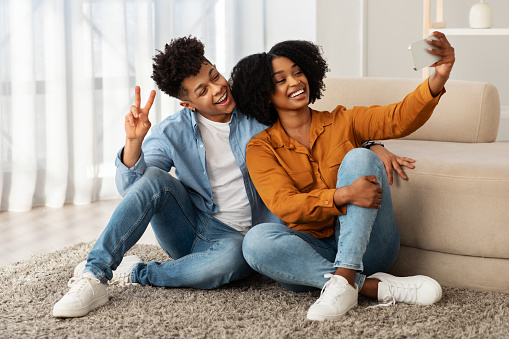 Sitting comfortably on the floor, a delighted young african american couple enjoys a light-hearted moment taking a selfie, with the man giving a peace sign and both wearing stylish denim