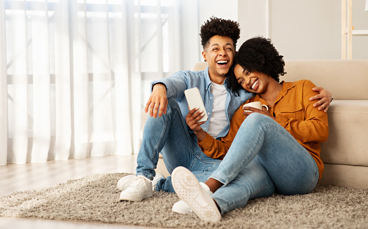 A joyful african american couple sitting on the floor against a couch, sharing a light-hearted moment with a smartphone, their laughter and closeness filling the room with warmth