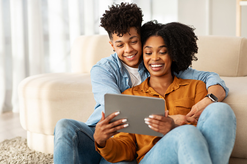 Snuggled up on the couch, african american young couple in casual wear shares a tender moment as they happily look at a tablet together in a cozy, light-filled living room