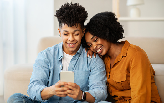 A joyous african american couple shares a warm, affectionate moment, leaning closely together as they view something amusing on their smartphone in a well-lit, cozy room