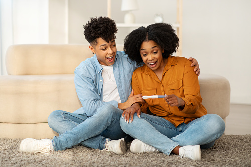 An overjoyed young african american couple sits on the floor with a positive pregnancy test, sharing an exhilarating moment of joy as they react to the good news in a cozy living room