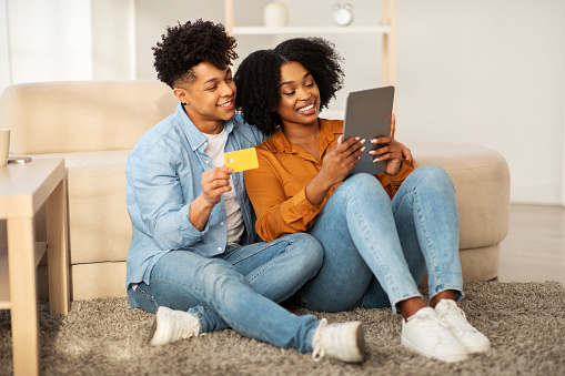 Snuggled on the floor against a couch, a young african american couple shares a digital tablet and a credit card, happily engaged in online shopping or planning in a homely environment