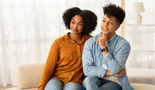 Glad African American millennial couple sits closely on sofa, woman and man is smiling thoughtfully, both enjoying a relaxed and comfortable moment together in a light-filled room