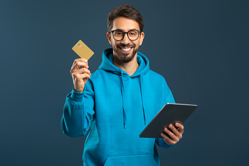 Online Payments. Cheerful Young Man Using Digital Tablet And Credit Card While Standing Against Blue Background In Studio, Smiling Millennial Guy Recommending App For E-Commerce, Copy Space