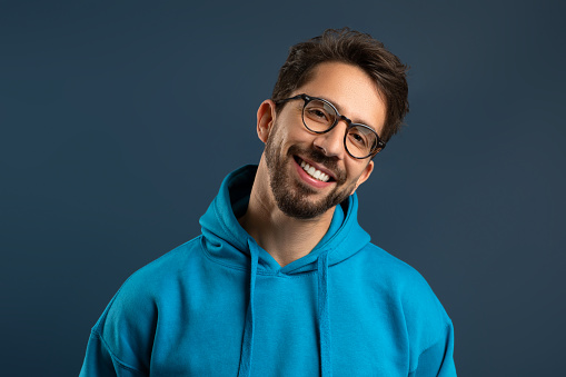 Smiling young man with stubble wearing glasses and bright hoodie standing against blue background, handsome millennial guy looking at camera while posing in studio, exuding casual confidence