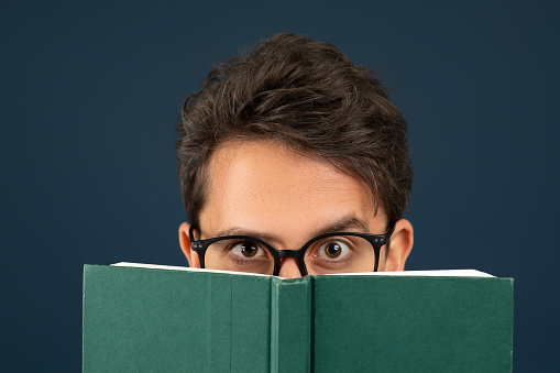 Young man wearing eyeglasses peeking over the top of green book, nerdy male showing curiosity and interest, standing isolated on dark blue studio background, enjoying reading, closeup