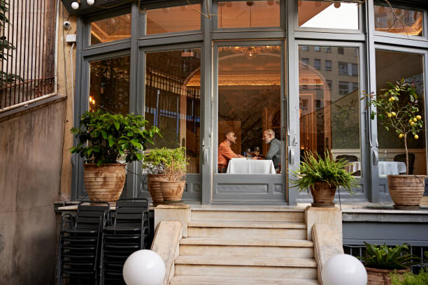 Men photographed through restaurant window during wine date Outdoor view of mature couple seated at table for two, face to face, enjoying drinks and conversation. gay dinner table stock pictures, royalty-free photos & images