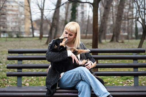 Young generation Z hipster girl sitting in park on a bench with her adopted dog, whom she rescued from the street from starvation, and gives him love and attention. Animal lover enjoying with her pet.