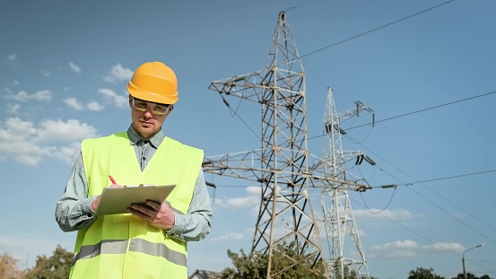 Energy auditor writing on clipboard and smiling at camera while standing near electricity pylons during site inspection. Dolly shot