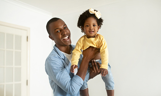 Happy young black father lifting up his pretty little daughter, joyful african american daddy and kid baby girl having fun together at home, looking at camera and smiling, panorama