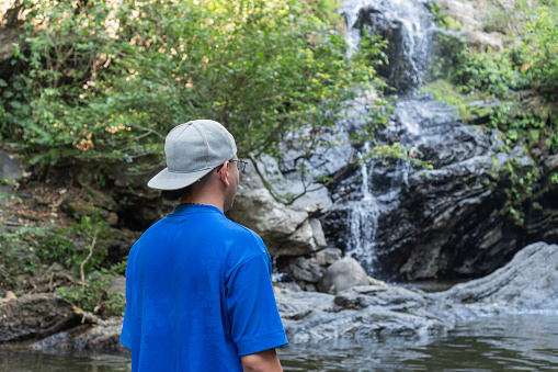 Man in blue standing in water, looking towards a sunlit forest waterfall.
