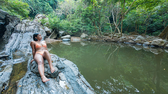 A thoughtful woman sits on a rock by a still pond in the forest, a waterfall in the distance.