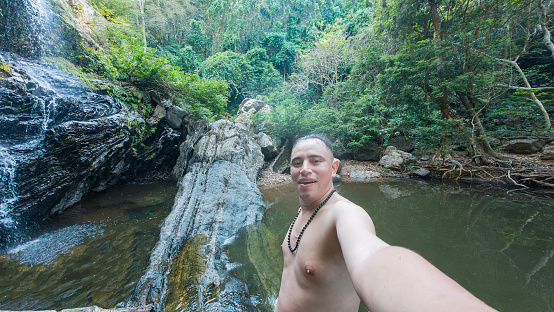 A man takes a selfie with a picturesque waterfall in a lush forest setting.