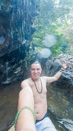 A person with a joyful expression sitting in water, reaching out to a waterfall.