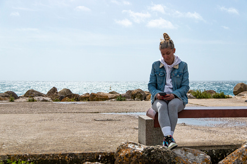 Blonde woman with bun using her mobile phone while enjoying the sea sitting on a bench on the promenade