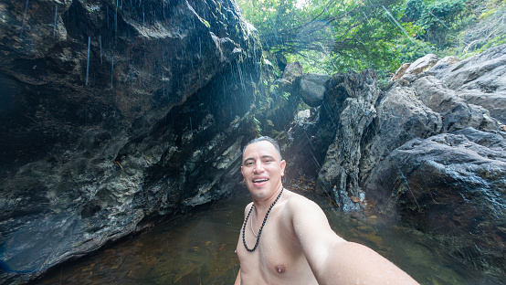 Smiling man captures a selfie while standing in a natural pool under a rocky waterfall surrounded by foliage.
