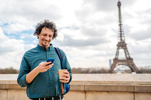 Handsome young man drinking coffee to go while using his smart phone in front of an Eiffel Tower from the street in Paris in France.